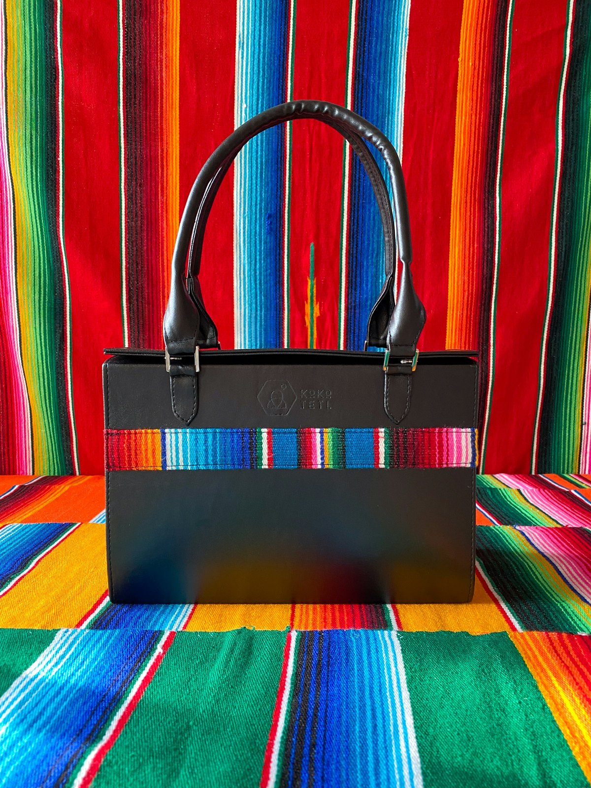 A black leather handbag with rolled handles standing on a brightly striped woven blanket, the same blanket hung behind it