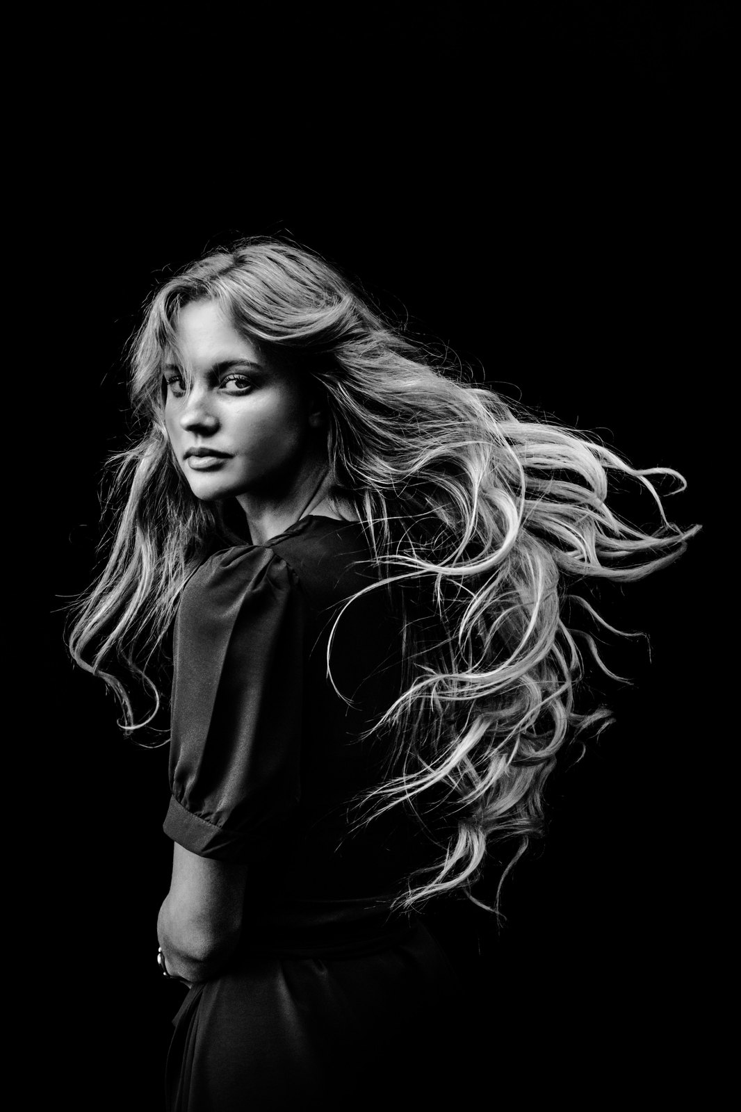 A black and white portrait of a woman with long hair flying, against a black studio backdrop